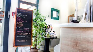 Un restaurante con un menú en pizarra que enumera varios platos y precios, una planta en maceta y un mostrador con libros y revistas.
