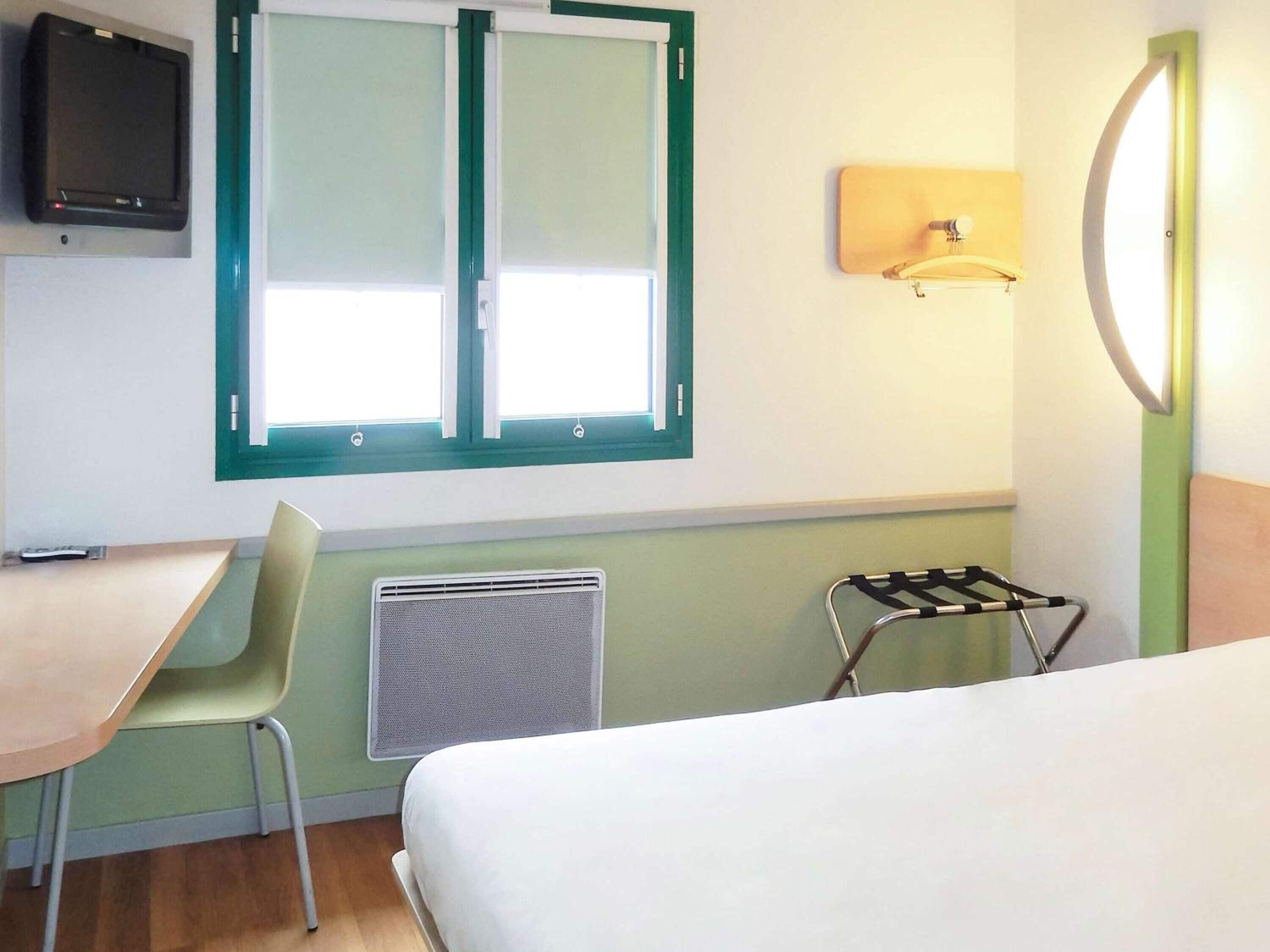 A hotel room with a bed, a desk, a chair, a radiator, a TV mounted on the wall, and a window with a view.