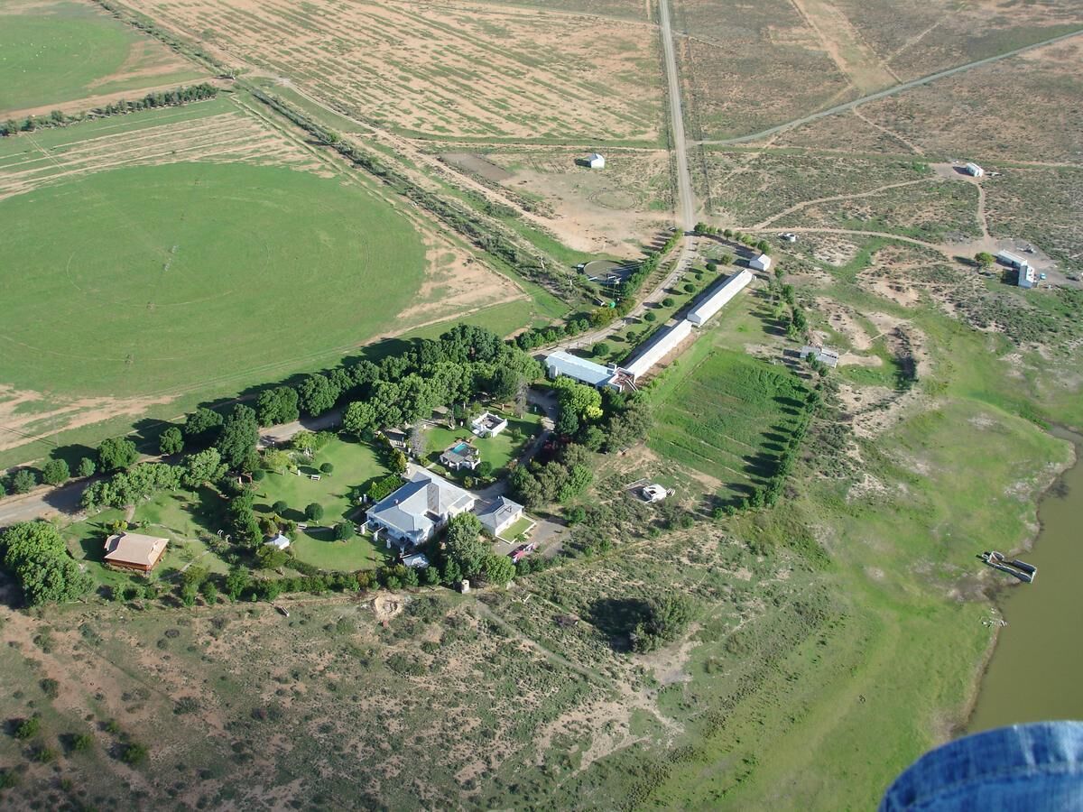 Une vue aérienne d’une propriété rurale comprenant un long bâtiment, plusieurs constructions plus petites, ainsi qu’un mélange de champs ouverts et de zones boisées.