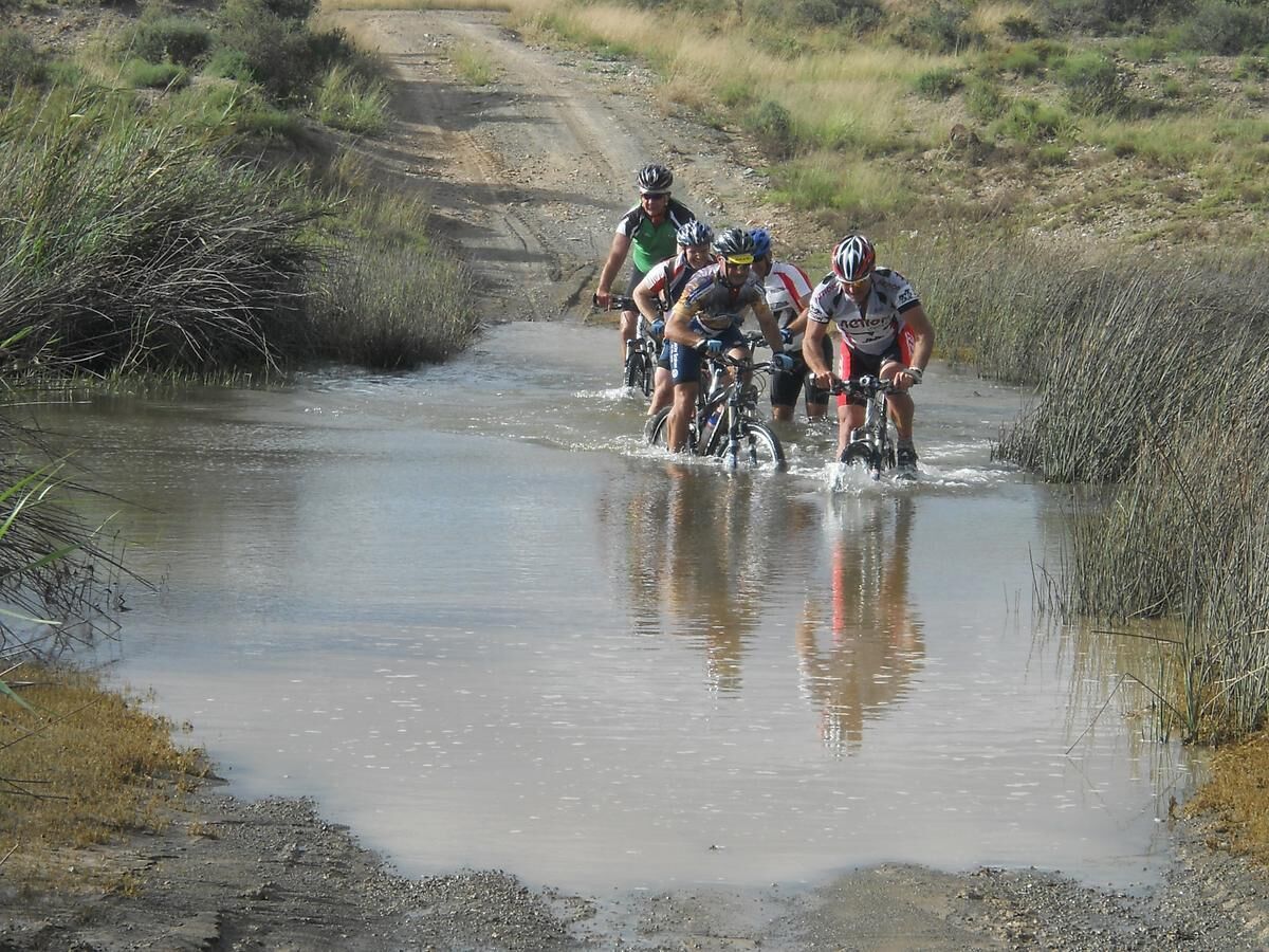Un grupo de ciclistas cruza un vado poco profundo en un camino de tierra rodeado de vegetación.