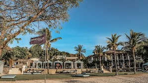 Un complex turístic a la vora de la platja amb una bandera que diu "PACIFICO Costa Rica". Hi ha gandules i ombrelles, un edifici amb un balcó i palmeres.