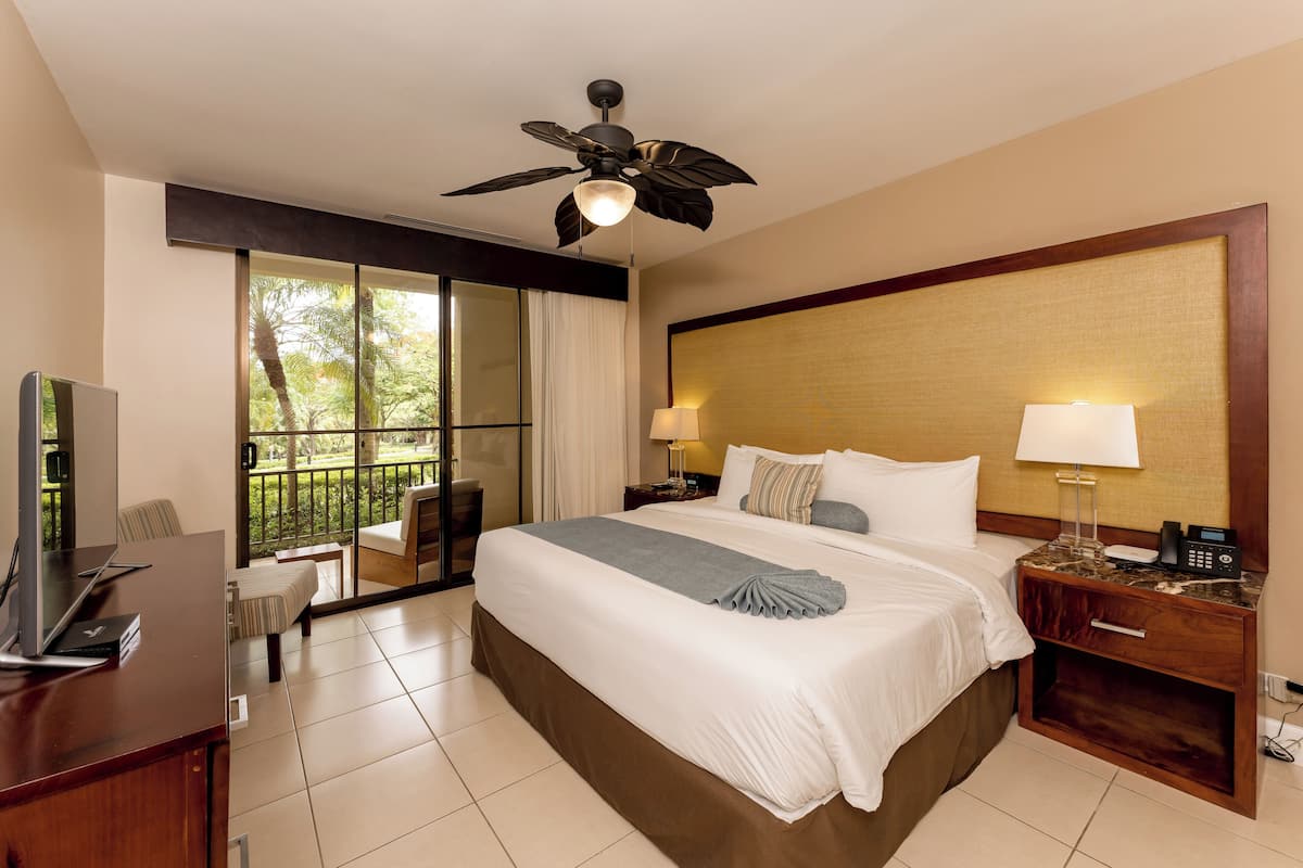 A hotel room with a large bed, a desk with a computer, a nightstand with a phone, and a ceiling fan.