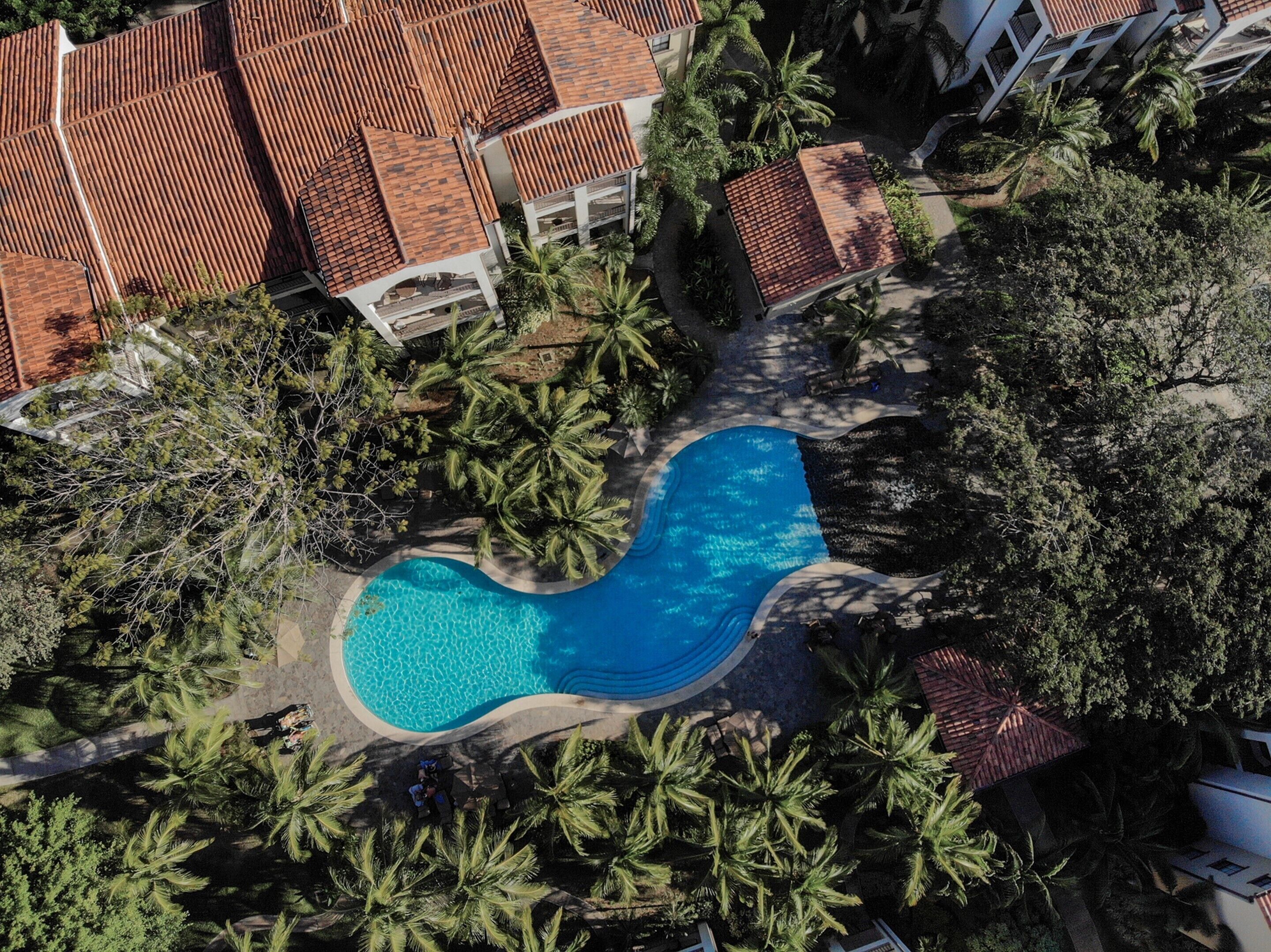 Vista aérea de un complejo turístico con piscina rodeada de palmeras y edificios de tejas rojas.