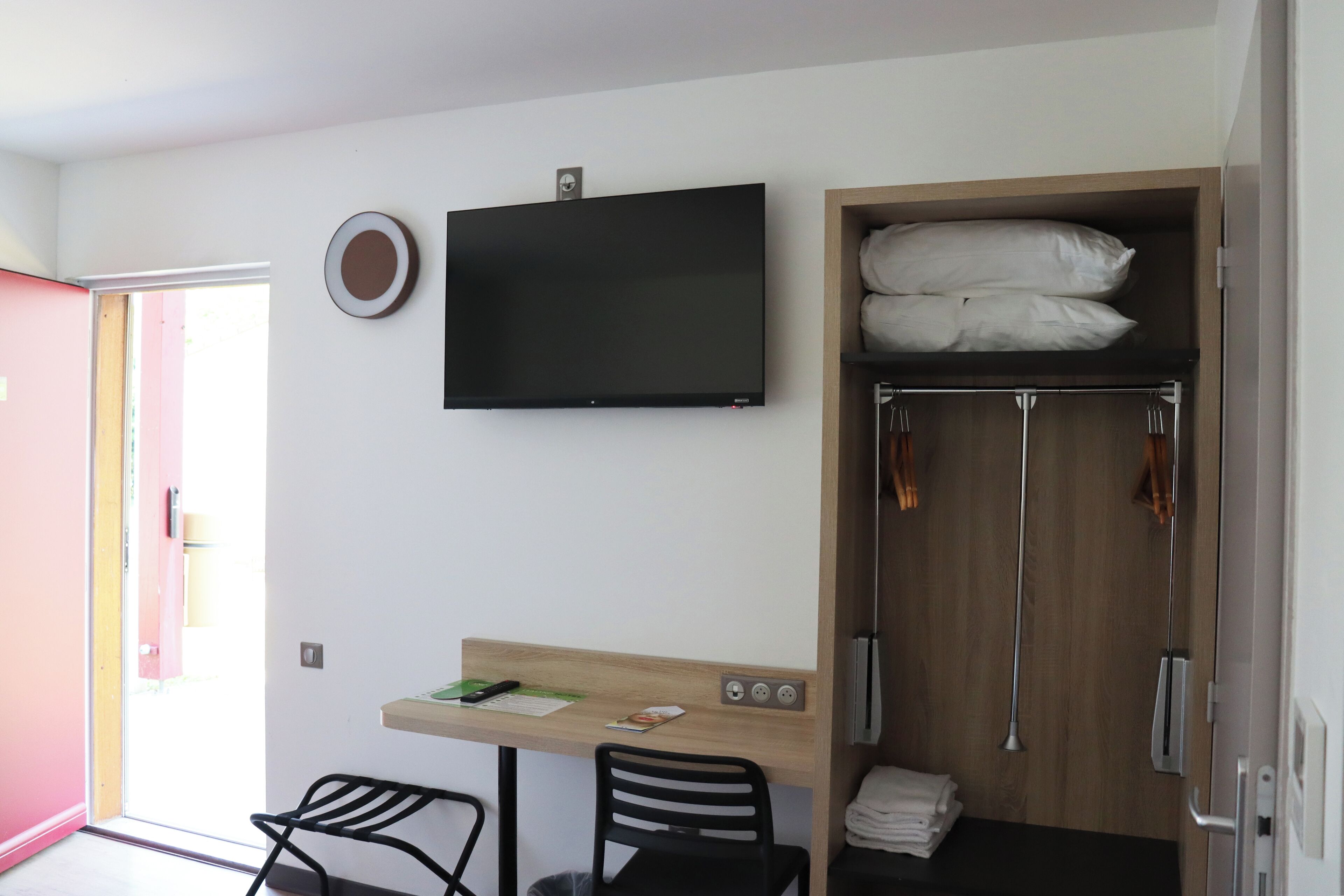 A small, modern room with a desk, two chairs, a TV, and a wardrobe with hanging clothes.