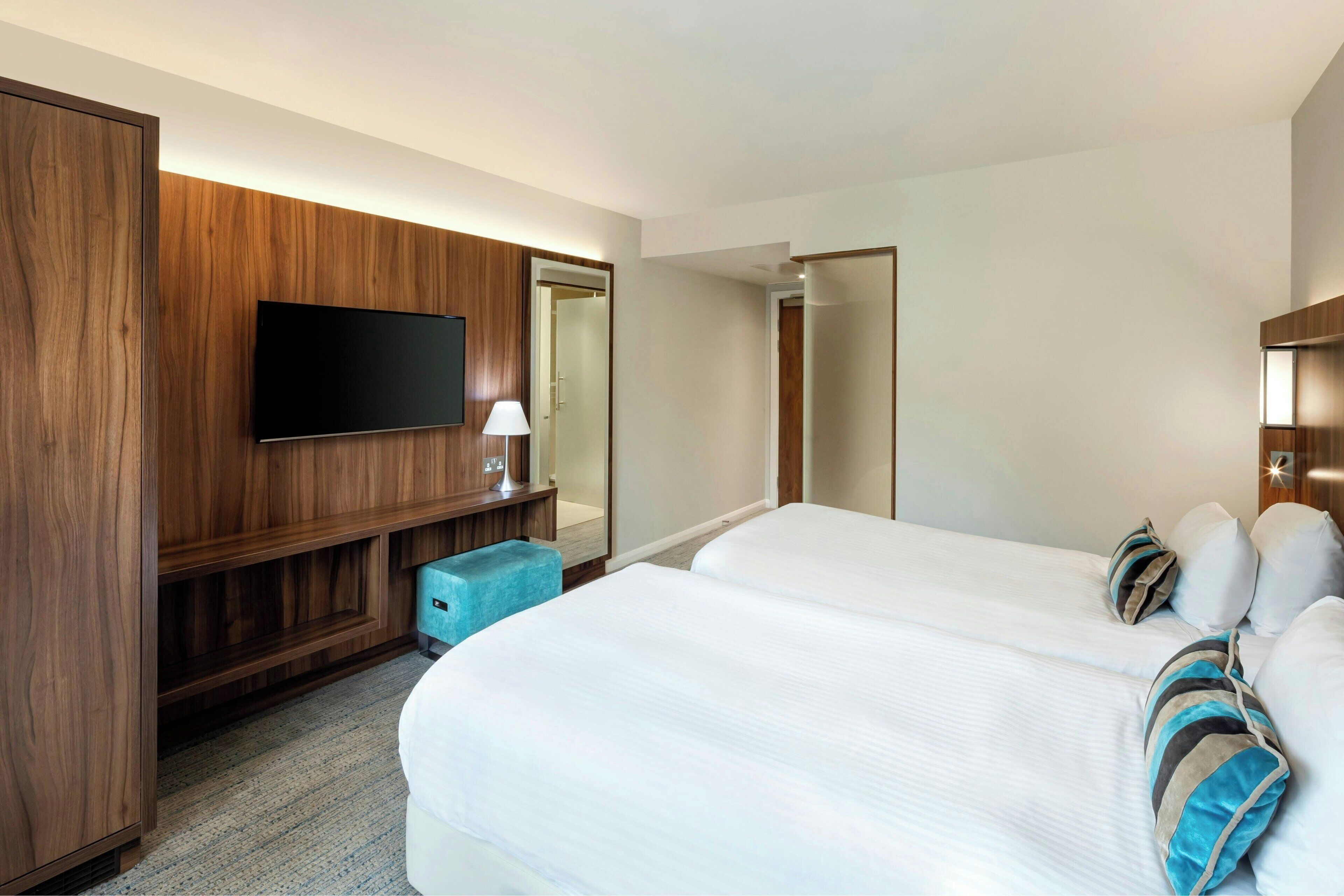 A hotel room with two beds, a TV mounted on a wooden panel, a small blue ottoman, and a mirror.