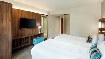 A hotel room with two beds, a TV mounted on a wooden panel, a small blue ottoman, and a mirror.