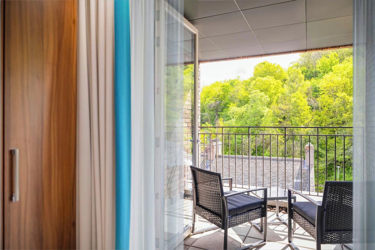 A balcony with a view of a forest, a glass door, and two wicker chairs.