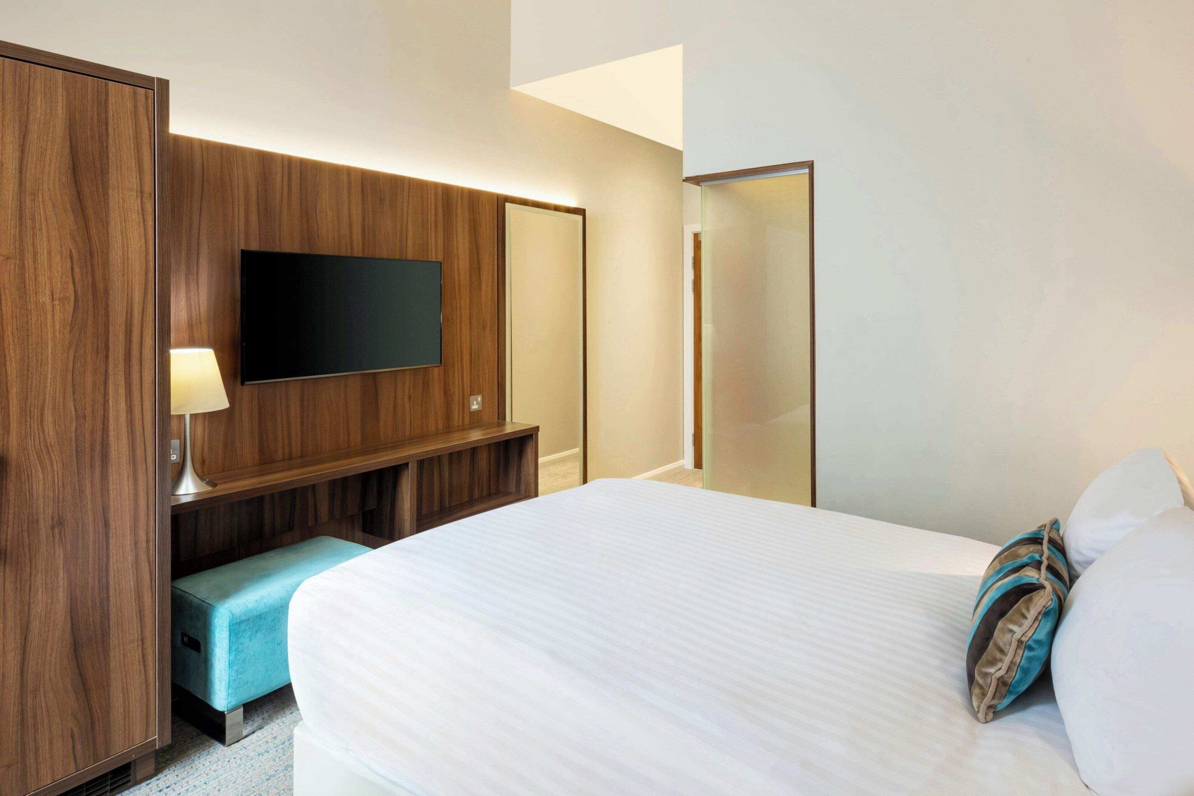 A hotel room with a bed, a TV mounted on a wooden panel, a bench, and a mirror.
