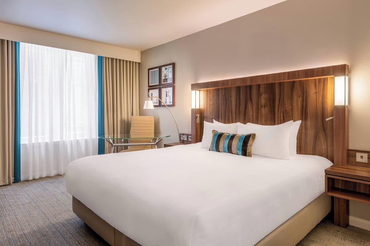 A hotel room with a large bed, a glass table, a chair, and a wooden headboard.