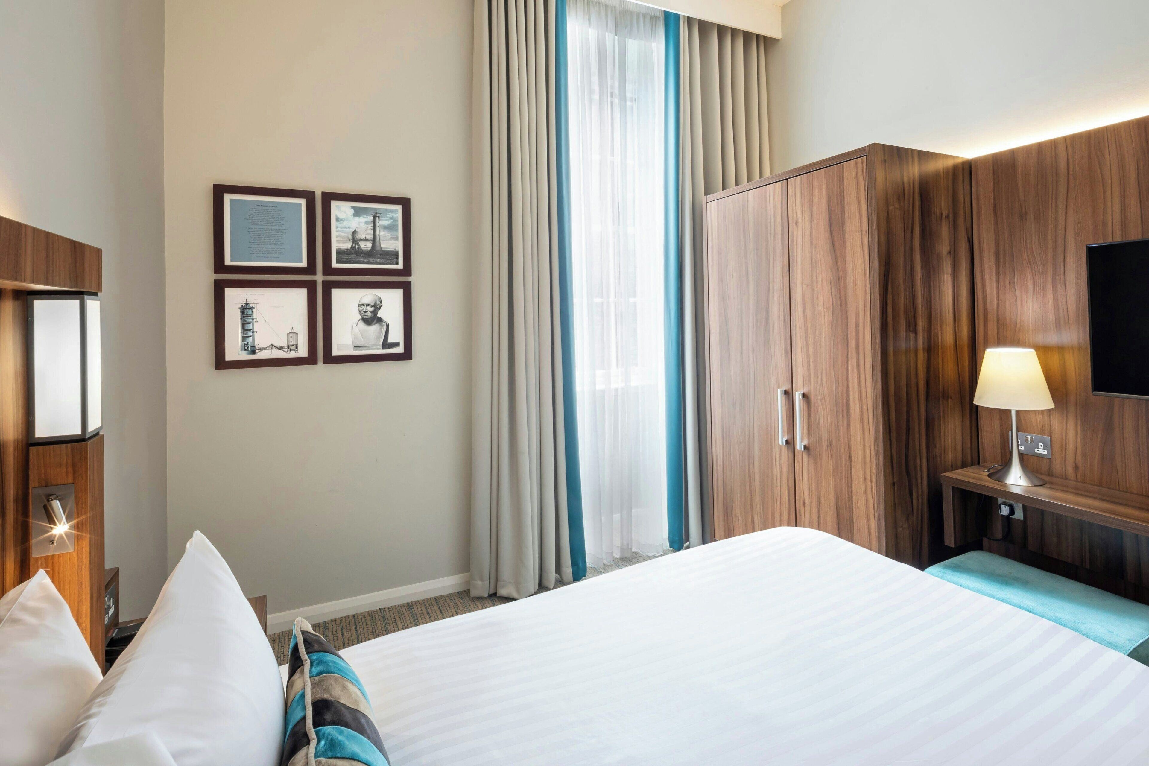 A hotel room with a large bed, a desk with a lamp, a TV mounted on the wall, and a set of framed pictures on the wall.