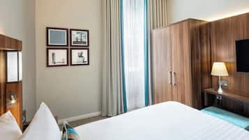 A hotel room with a large bed, a desk with a lamp, a TV mounted on the wall, and a set of framed pictures on the wall.