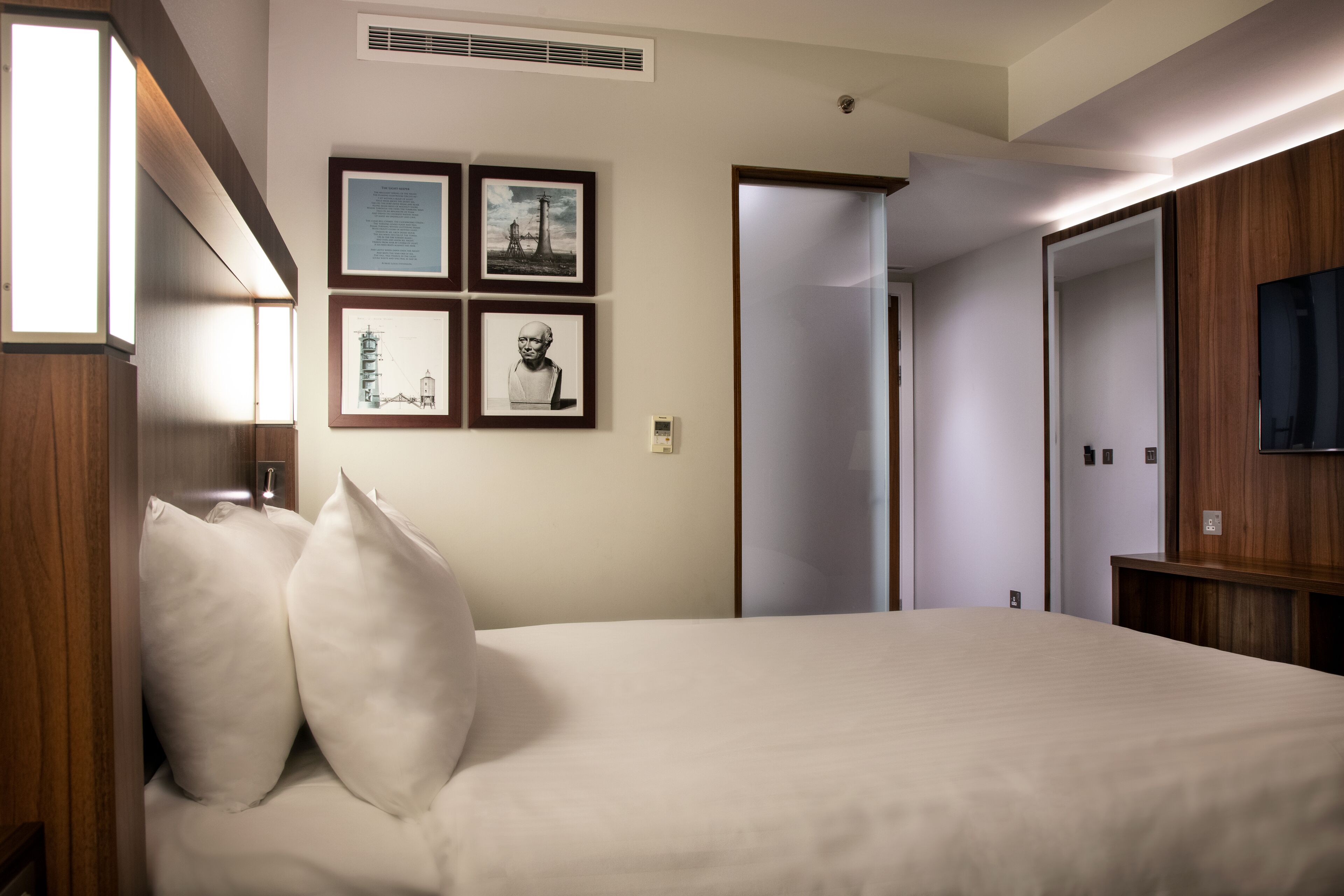A hotel room with a bed, pillows, a flat-screen TV, and framed pictures on the wall.