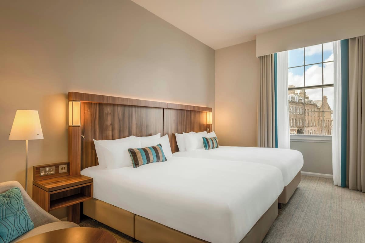 A hotel room with a large bed, a bedside table with a lamp, and a view of a cityscape through a window.