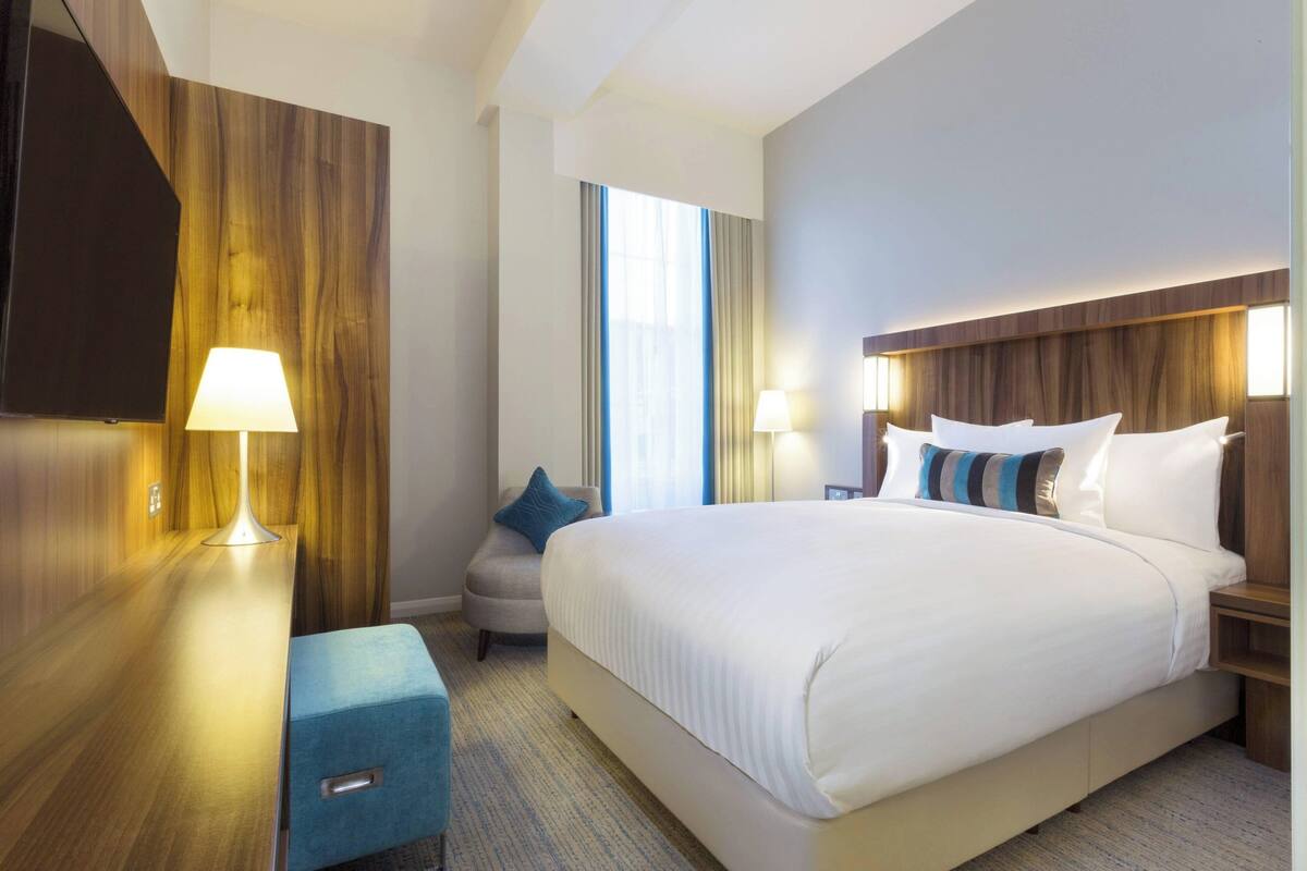 A hotel room with a large bed, bedside lamps, a desk with a lamp, a chair, and a TV on the wall.