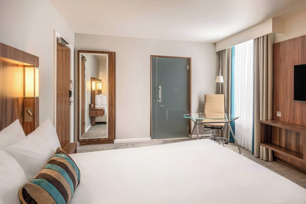 A hotel room with a large bed, a glass table, a chair, a TV, and a door leading to a bathroom.