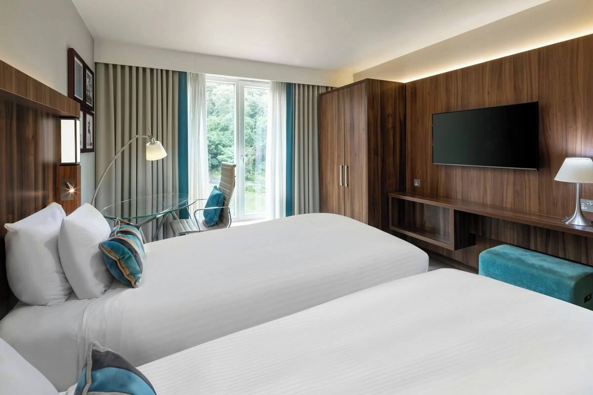 A hotel room with two beds, a TV mounted on the wall, a desk with a lamp, and a chair near a window with curtains.