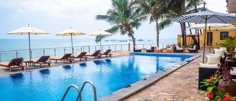 A pool area with lounge chairs and umbrellas, a stone-tiled deck, and a view of the ocean.