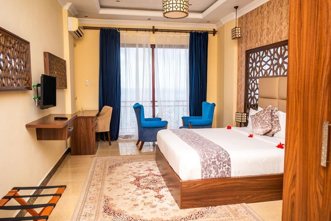 A hotel room with a bed, two blue chairs, a desk, a TV, and a balcony with curtains.
