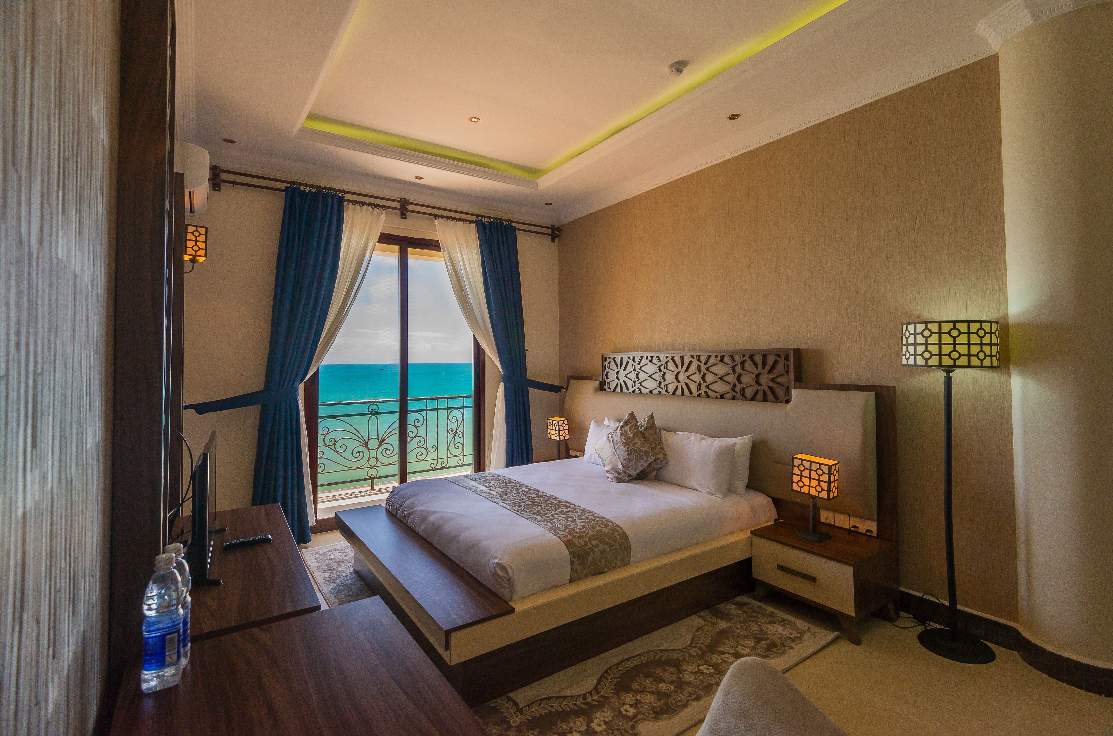 A hotel room with a bed, bedside tables, a desk, a lamp, and a view of the sea.