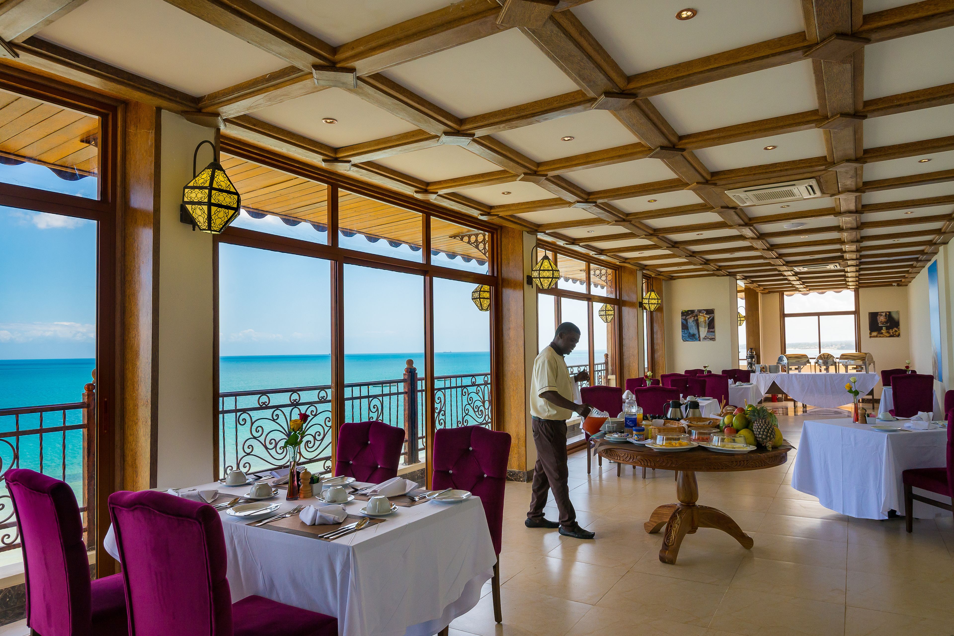 Un restaurante con mesas preparadas para comer, un camarero atendiendo a los clientes y vistas al océano a través de amplias ventanas.