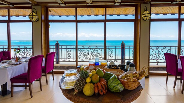 Comedor con vista al océano, con una mesa de buffet con frutas y pan, y sillas moradas.