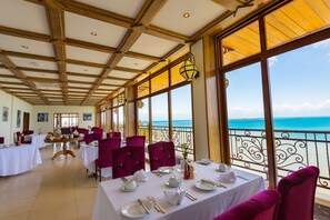 Un restaurant avec des tables dressées pour le service, de grandes fenêtres offrant une vue sur la mer et un plafond en bois.