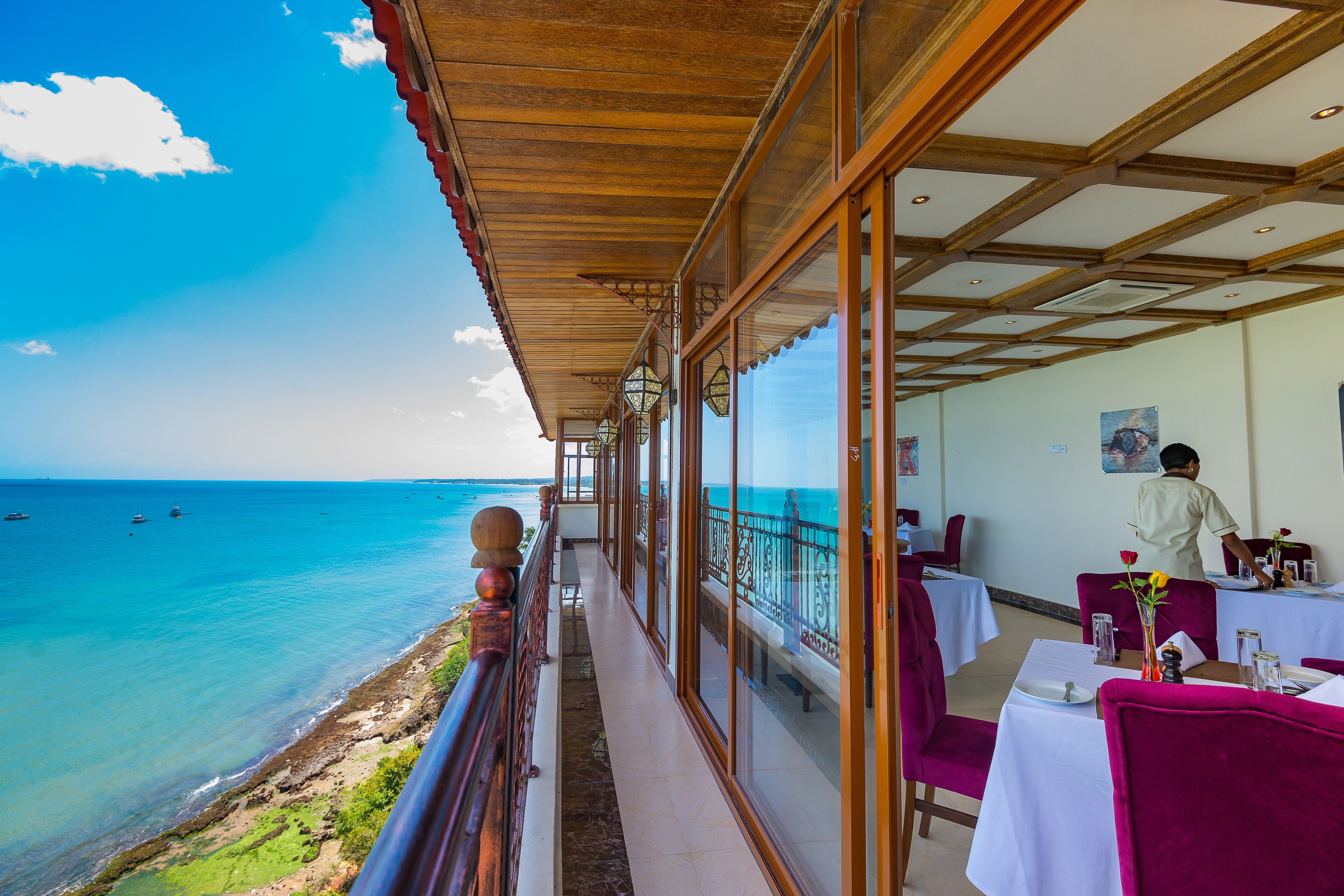 A seaside restaurant with a balcony overlooking the ocean, tables set with white tablecloths, and a view of boats on the water.