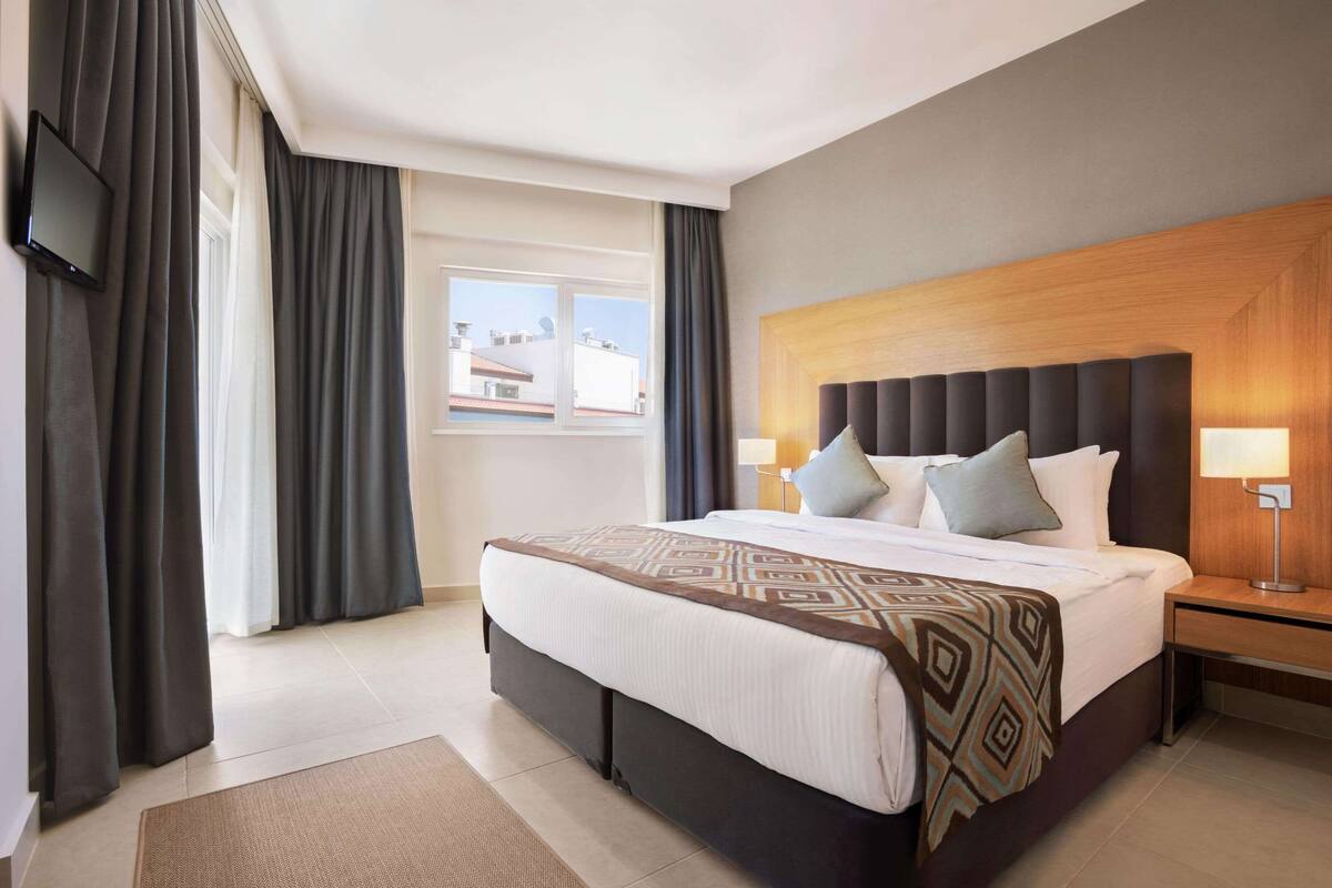 A hotel room with a large bed, a wooden headboard, two bedside tables with lamps, a flat-screen TV mounted on the wall, and a view of buildings through the window.