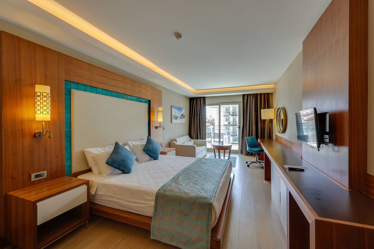 A hotel room with a bed, bedside tables, a desk, a chair, a TV, and a window with curtains.
