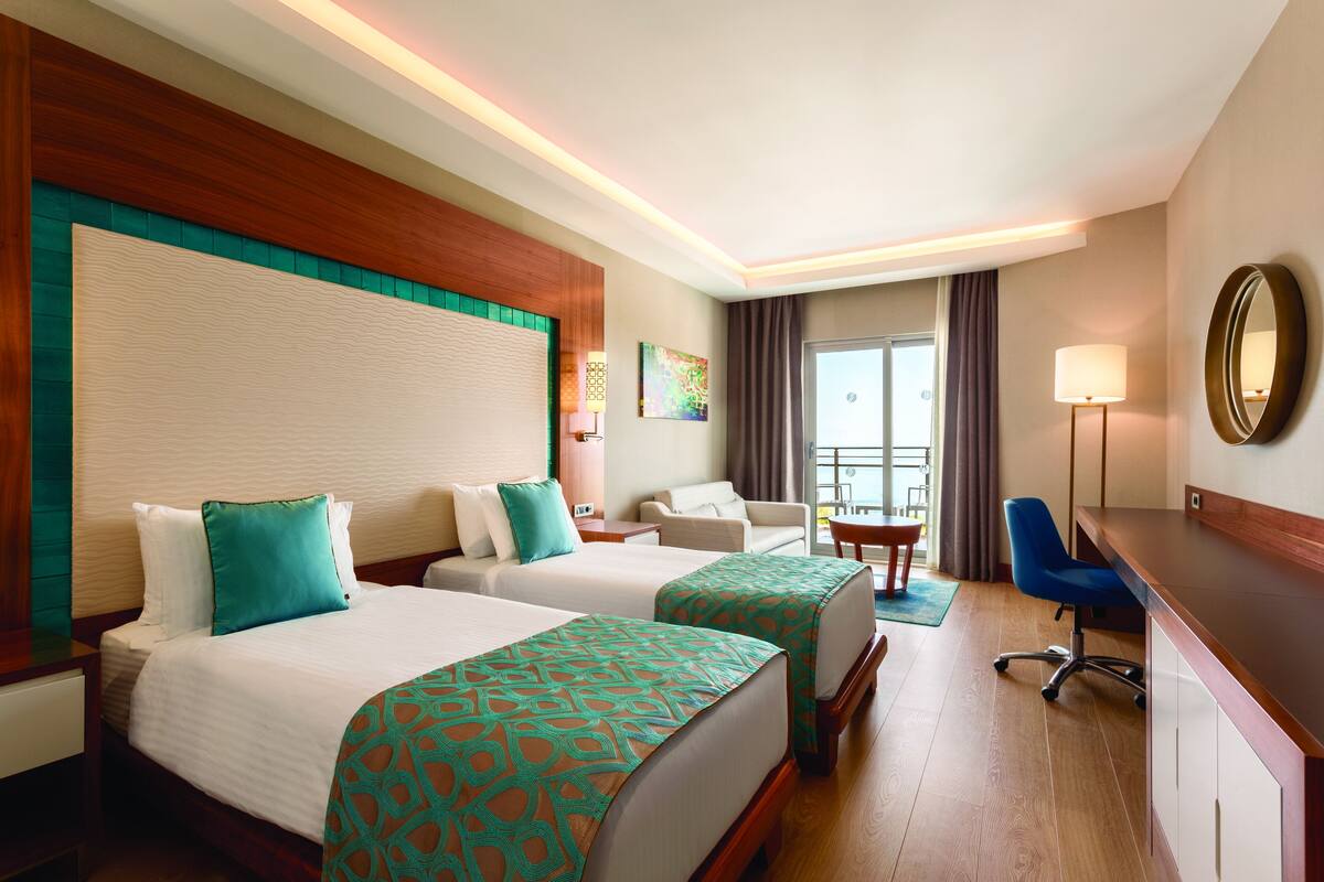 A hotel room with two beds, a desk, a chair, a sofa, and a balcony with curtains.
