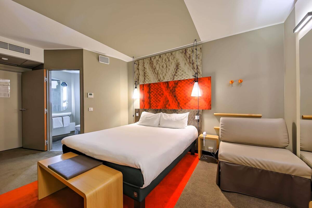 A hotel room with a bed, a desk, a chair, a red carpet, and a bathroom visible through an open door.
