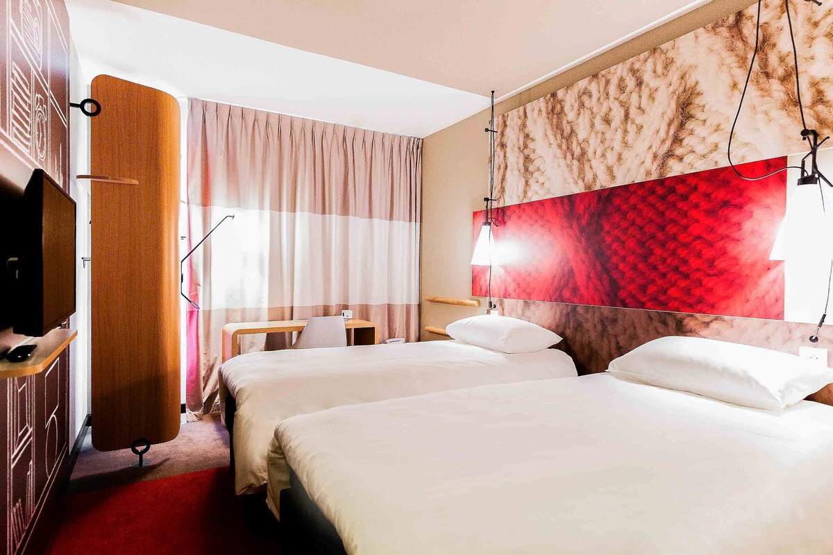 Habitación de hotel con dos camas, un escritorio, un televisor y un mural grande de color rojo y beige.