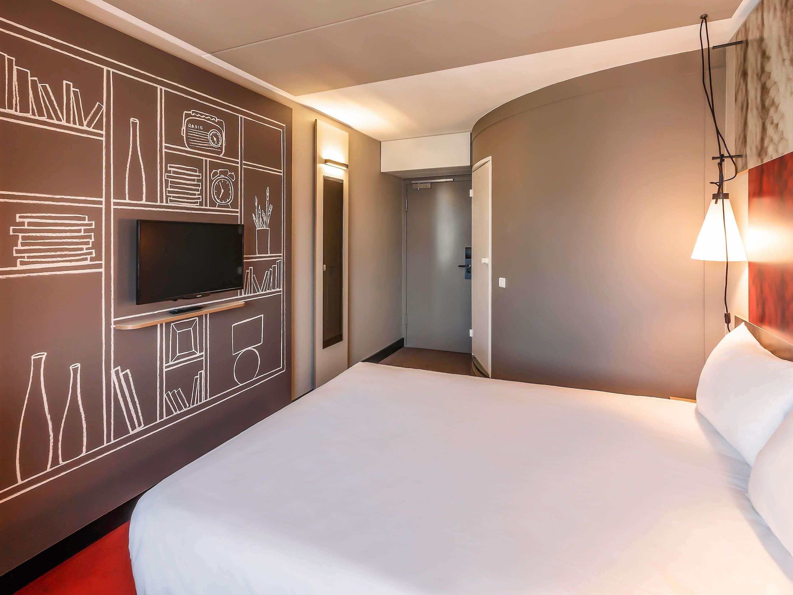 A hotel room with a large bed, a flat-screen TV mounted on the wall, and a bedside lamp.