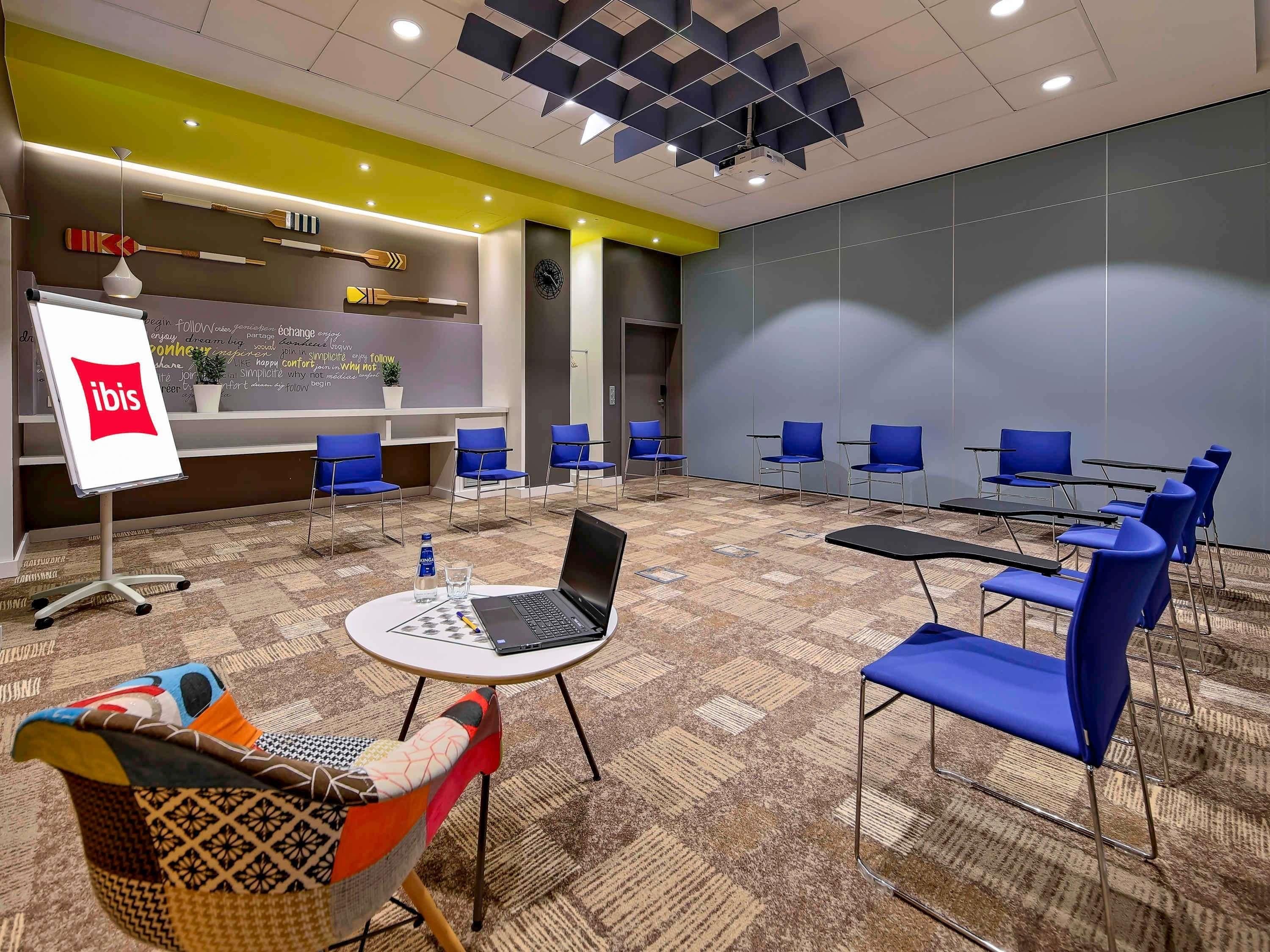 A conference room with blue chairs, a laptop on a table, and a whiteboard with 'ibis' logo.