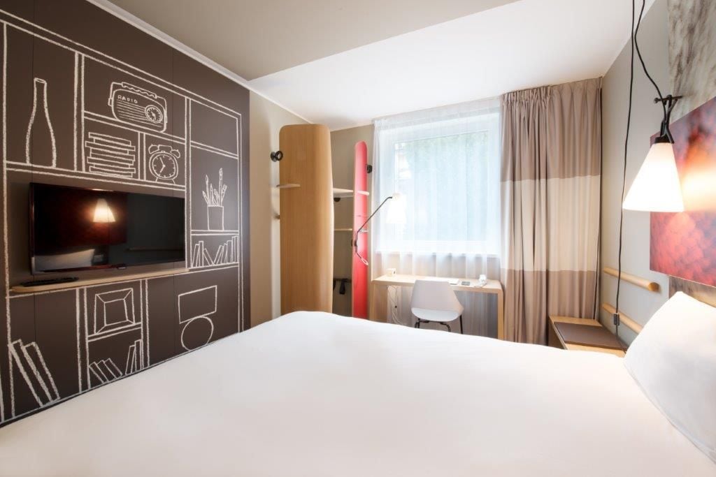 A hotel room with a bed, a desk, a chair, a television, and a surfboard.