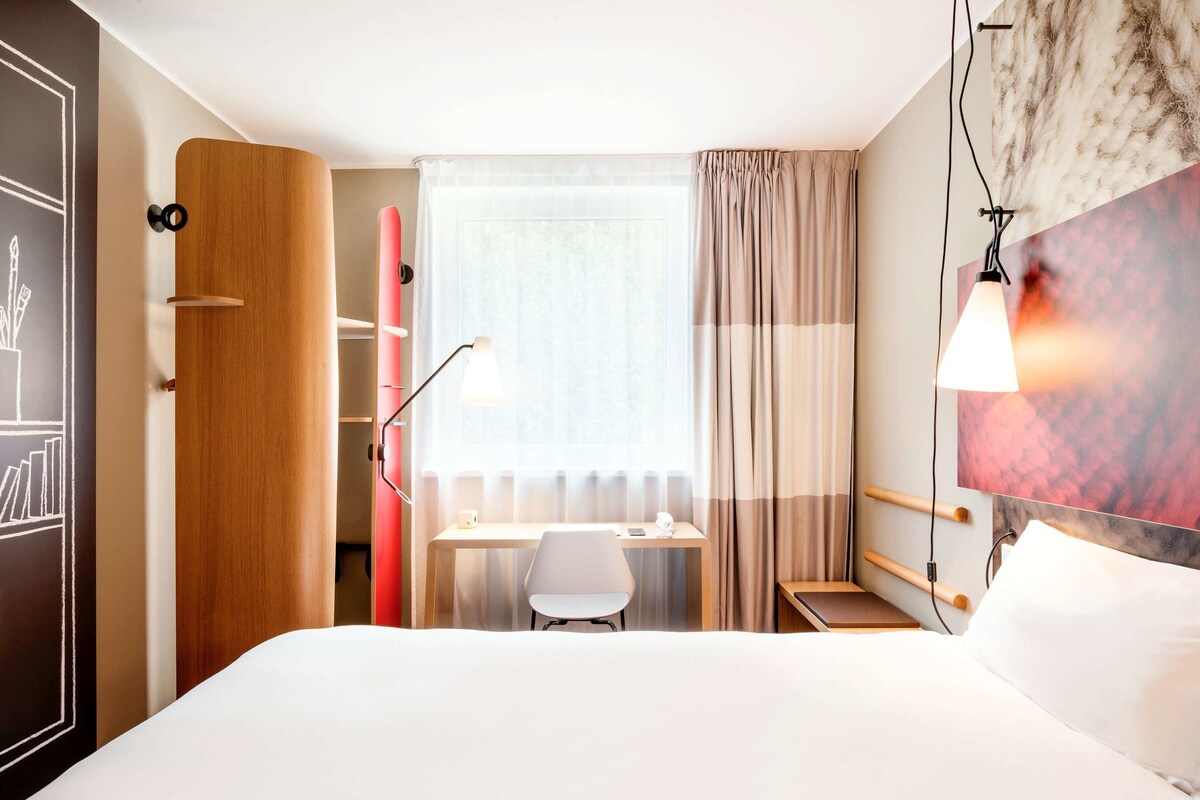 Habitación de hotel con cama, escritorio, silla, armario y ventana con cortinas.