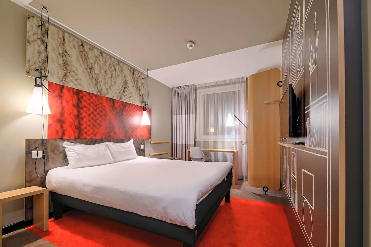 A hotel room with a bed, bedside tables, a desk, a chair, a TV, and a wardrobe.
