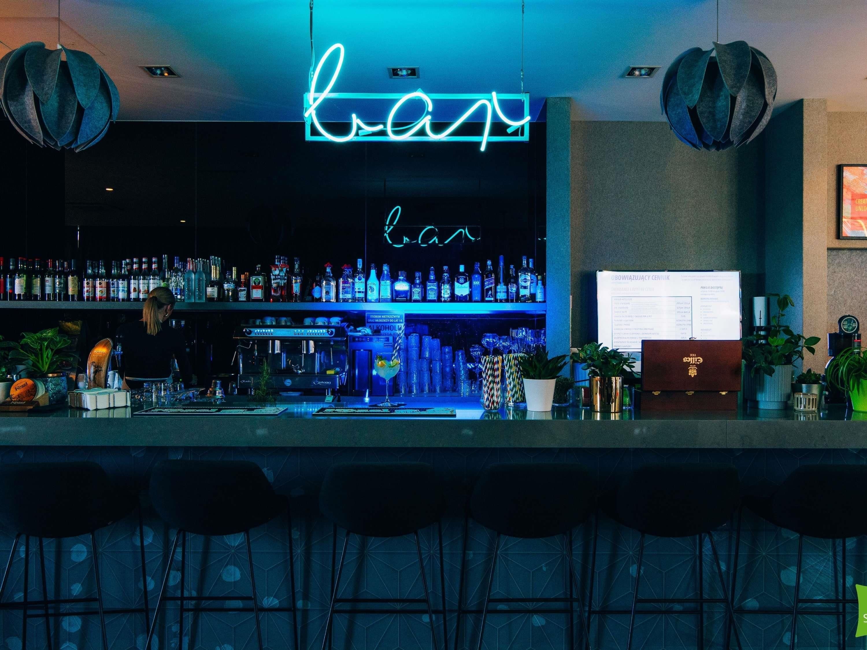 Zona de bar amb un senyal neó, amples prestatges plens de garrafes de licors, un panell amb el menú i tamborets de bar.