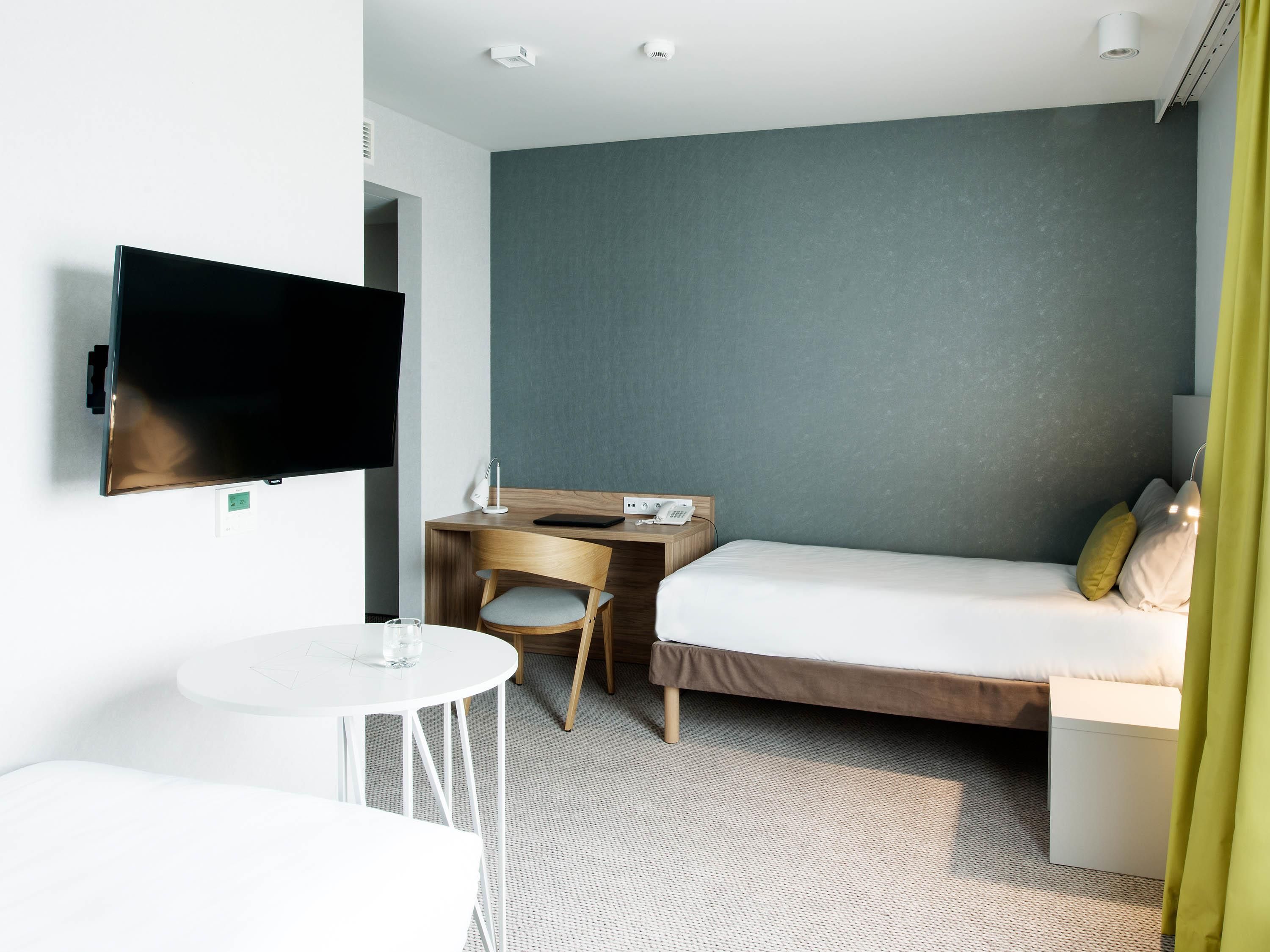 A hotel room with a bed, a desk with a chair, a television mounted on the wall, and a small round table with a glass.