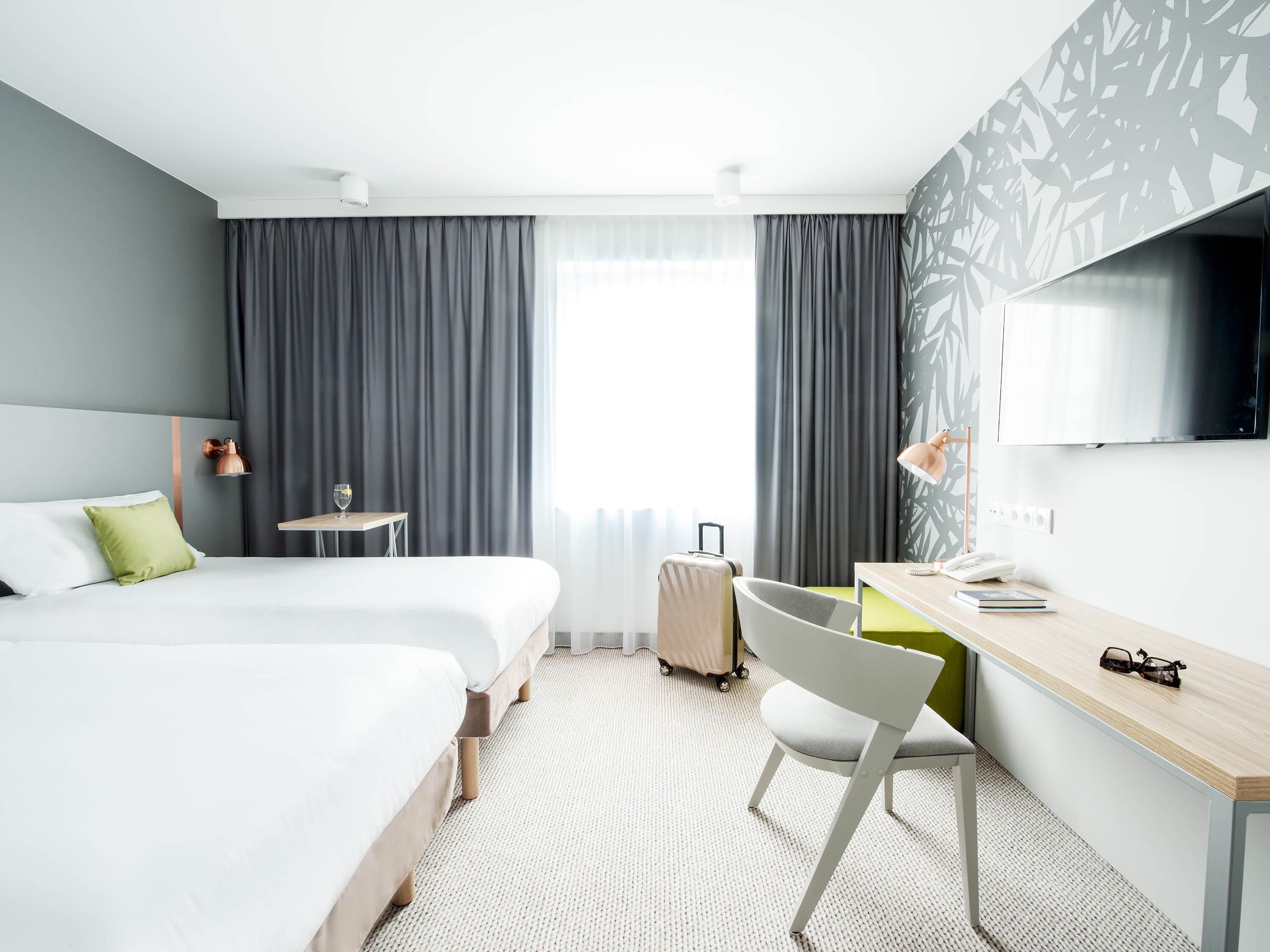A hotel room with two beds, a desk, a chair, a TV, and a suitcase.