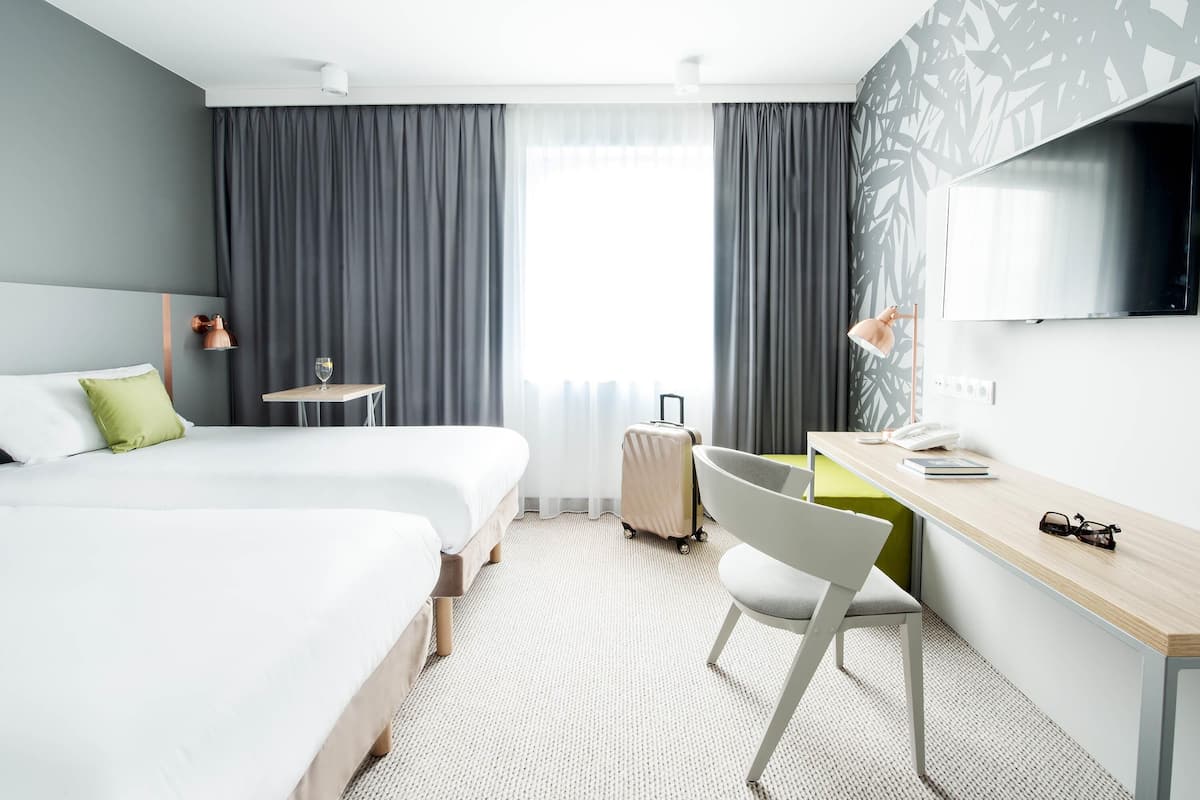 A hotel room with two beds, a desk, a chair, a TV, and a suitcase.