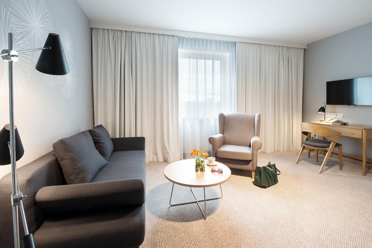 A modern hotel room with a sofa, armchair, desk, and TV.