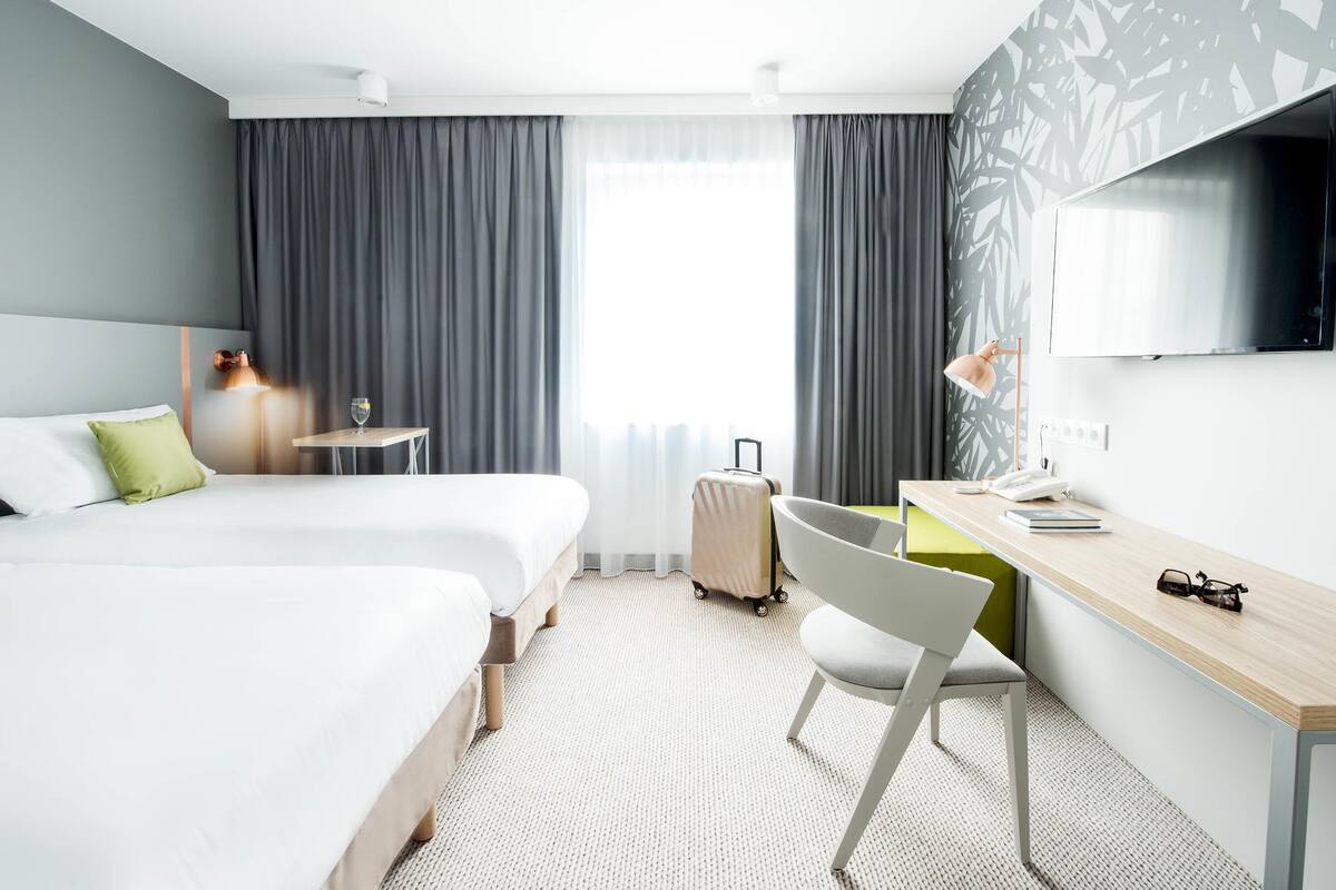 A modern hotel room with two beds, a desk, a chair, a TV, and a window with curtains.