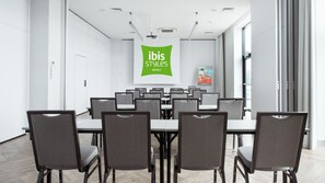 Una sala de conferencias con una gran mesa y sillas, un cartel verde de ibis Styles Hotels y un póster en la pared.