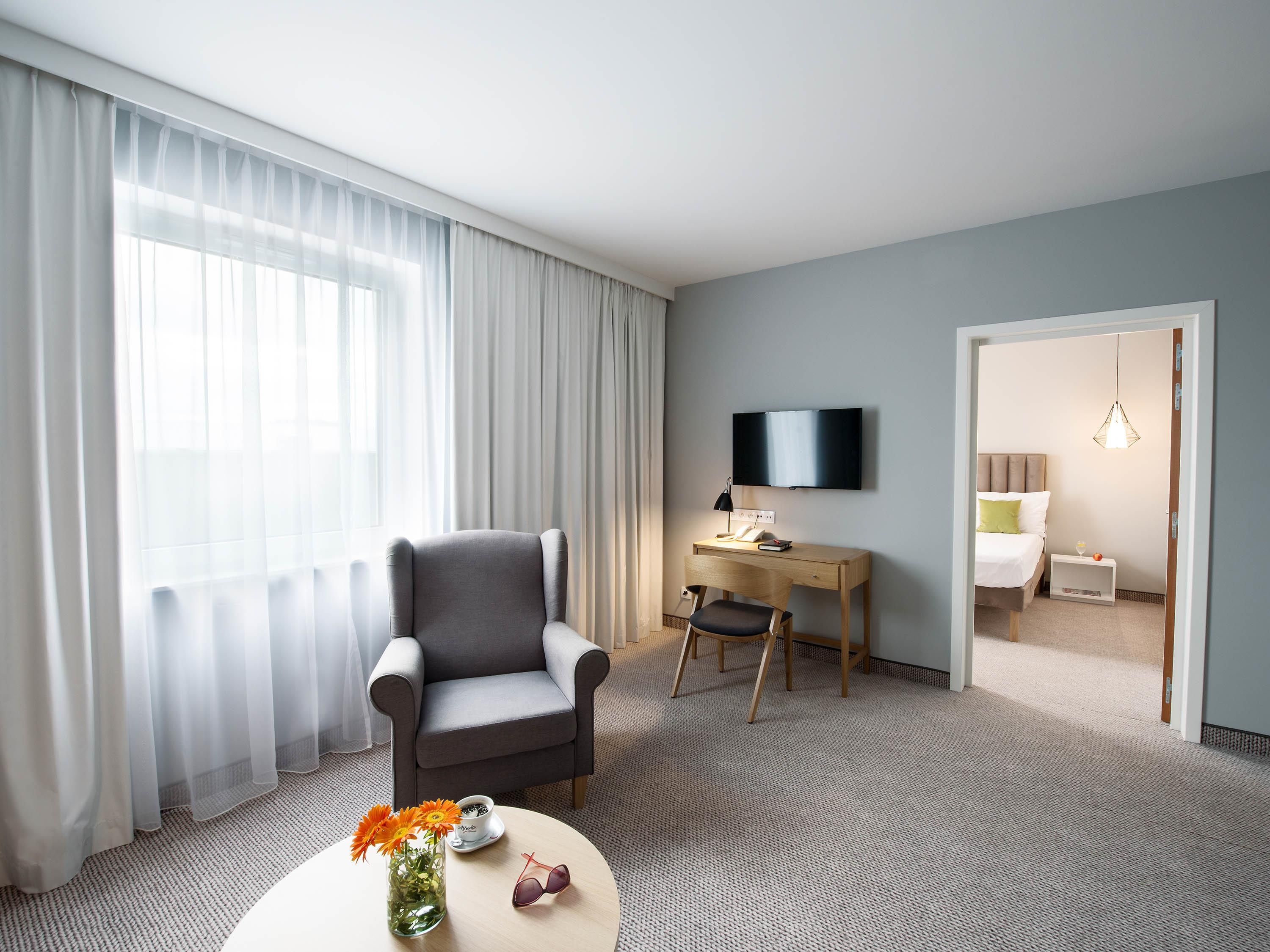 A hotel room with a grey armchair, a desk with a flat-screen TV, a bed with a headboard, and a small table with a vase of flowers and sunglasses.