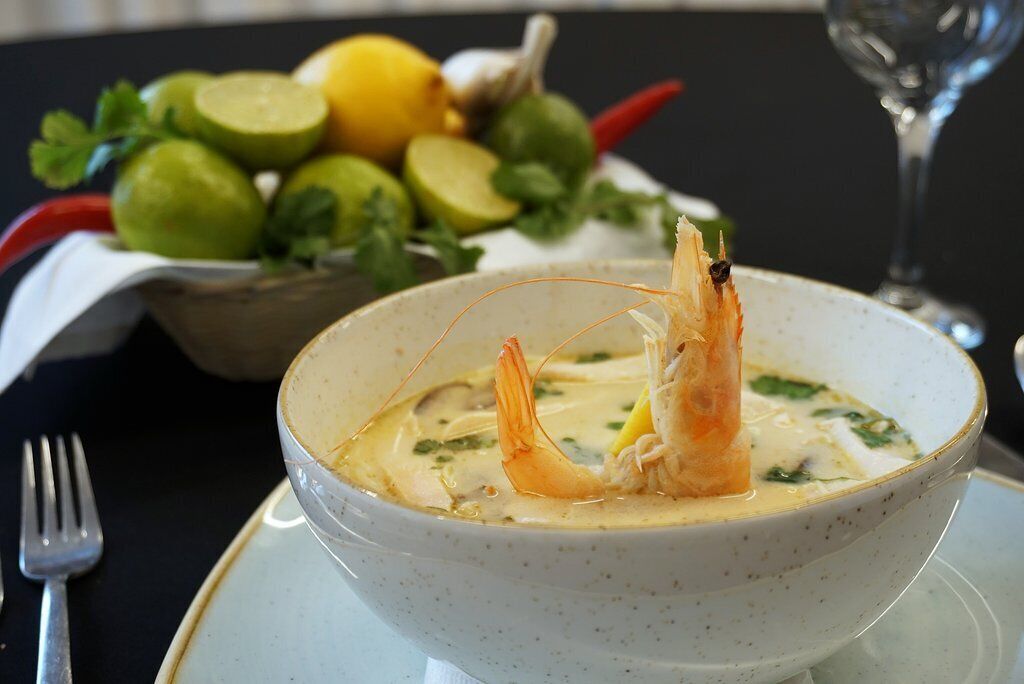 Un tazón de sopa de camarones, adornado con hierbas, y una cesta de verduras al fondo.