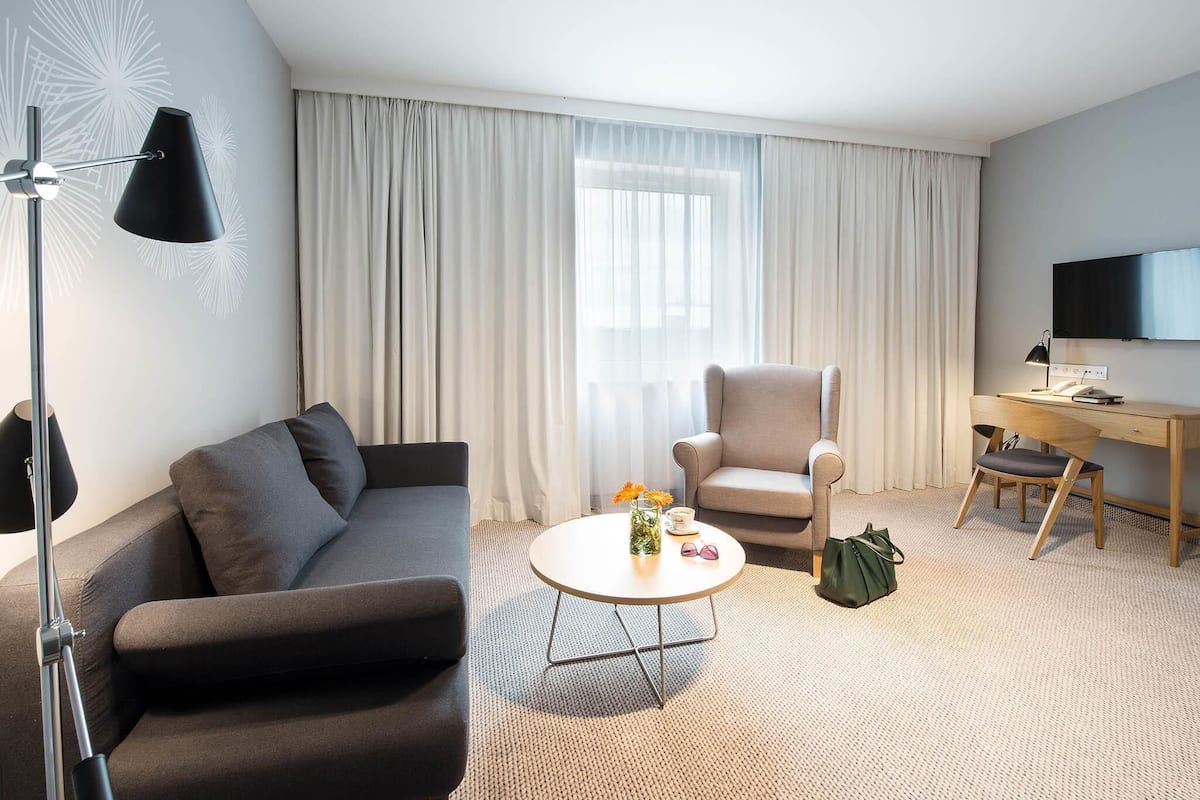 Ein modernes Hotelzimmer mit Sofa, Sessel, kleinem runden Tisch, Schreibtisch und Fernseher.