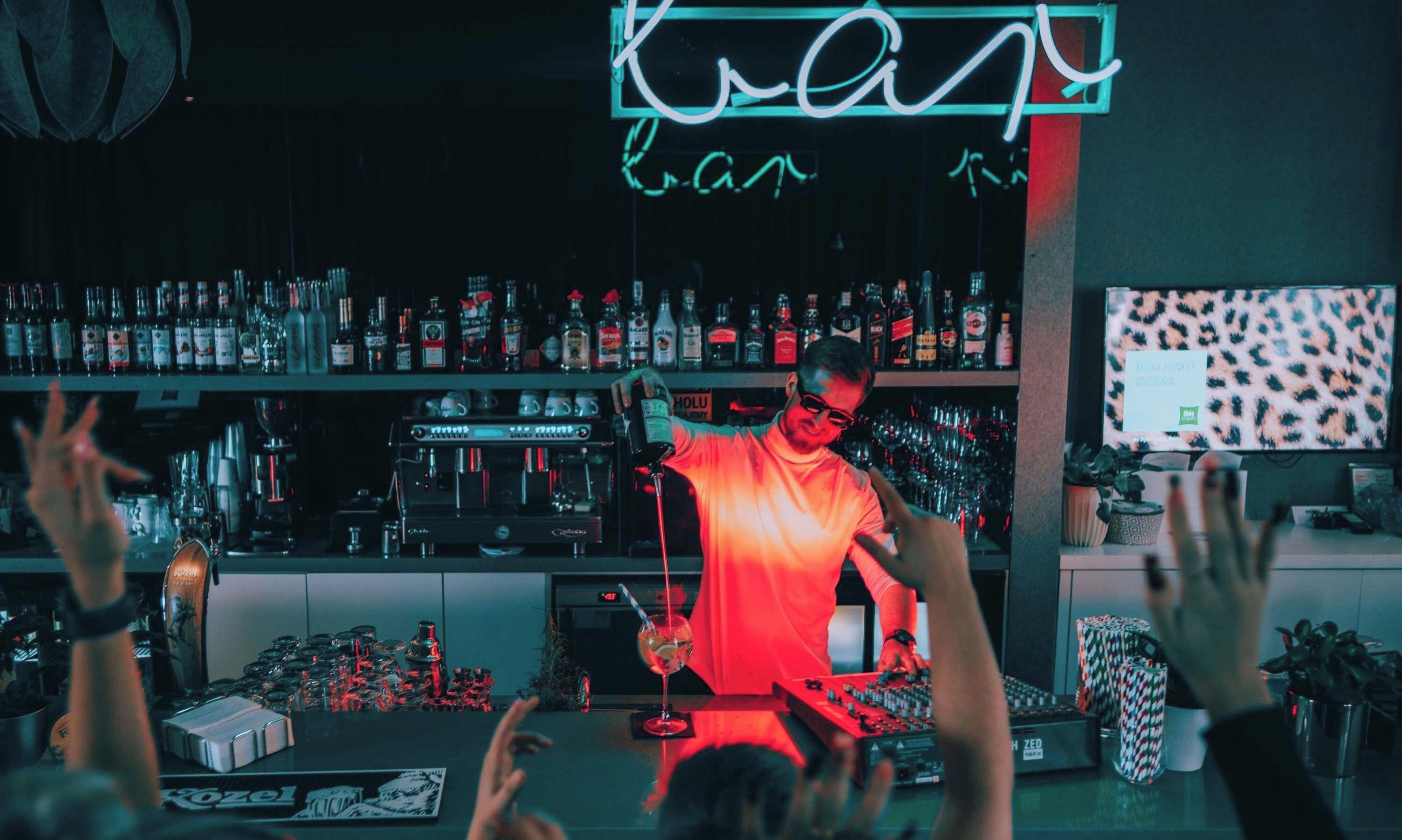 A bar with a neon sign, shelves of liquor bottles, a coffee machine, and a DJ booth.