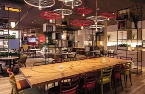 A modern interior with a central wooden table surrounded by colorful chairs, overhead circular light fixtures, and a variety of shelving units. - Leonardo Hotel Braunschweig City Center (Braunschweig)
