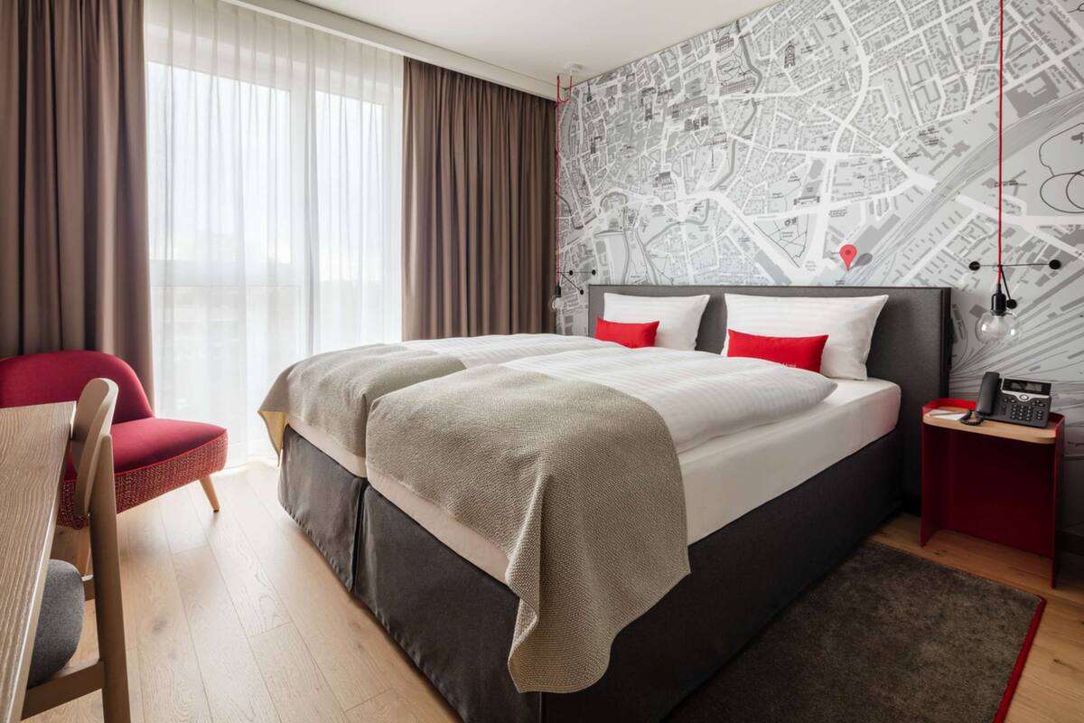 Quarto de hotel com uma cama grande, uma cadeira vermelha, uma escrivaninha e uma parede com mapa.